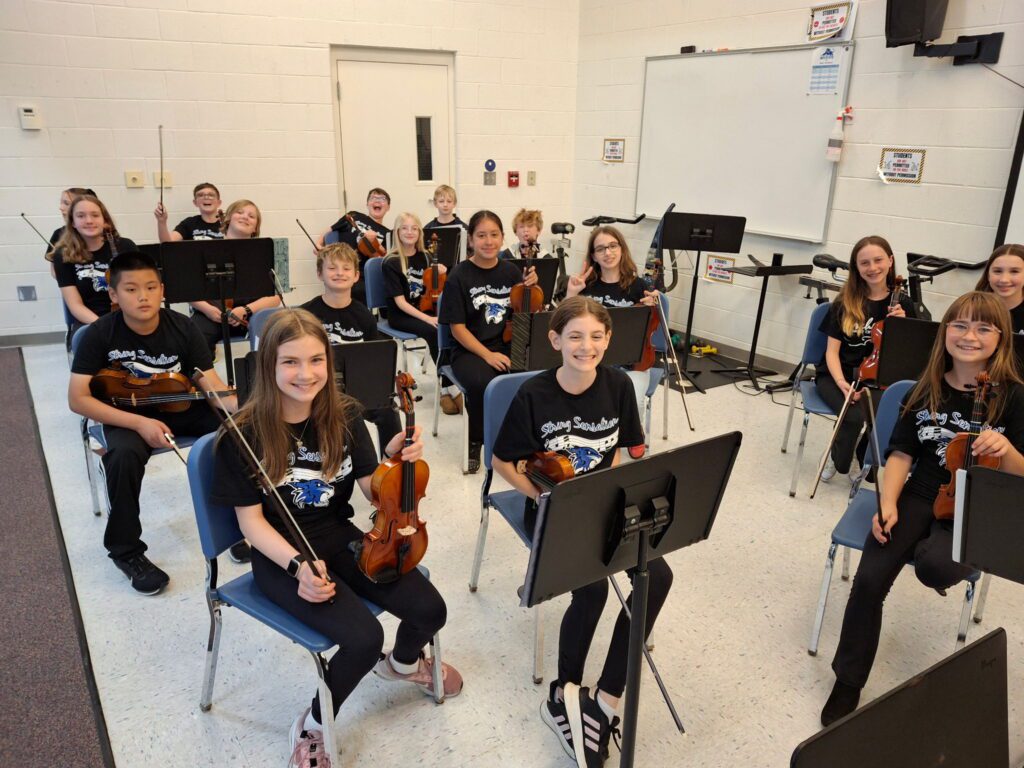 A group of violin and viola students holding their instruments and smiling in the Tharp gymnasium