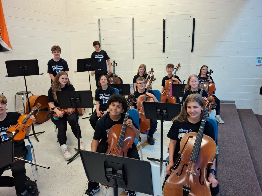 A group of cello, viola, and bass students holding their instruments and smiling in the Tharp gymnasium
